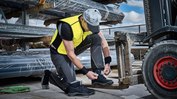 Bouwvakker met geel koelvest strikt zijn werkschoenen naast een heftruck op een zonovergoten bouwplaats.
