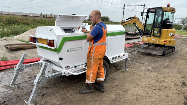 Haspelwagen en minigraver bij kabelwerkzaamheden op terrein met spoorlijnen op de achtergrond.