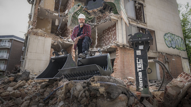 Demolition worker in a hard hat stands on a front-end loader bucket amid collapsed brick and rubble in front of a damaged building.