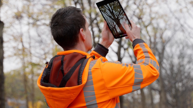Worker in a high-visibility orange jacket uses a tablet outdoors among trees, viewed from behind.
