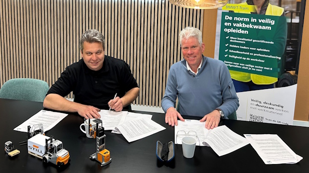Two people sit at a table signing documents, with small toy trucks and a mug on the desk and a promotional poster in the background.