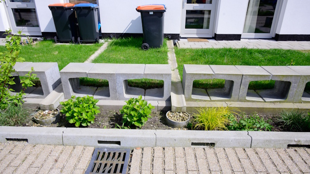 Concrete block planter with small shrubs and grasses in front of a modern white building; three trash bins behind.