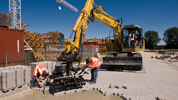 Construction workers in safety vests lay gray concrete pavers on a road while a yellow excavator helps position the blocks.