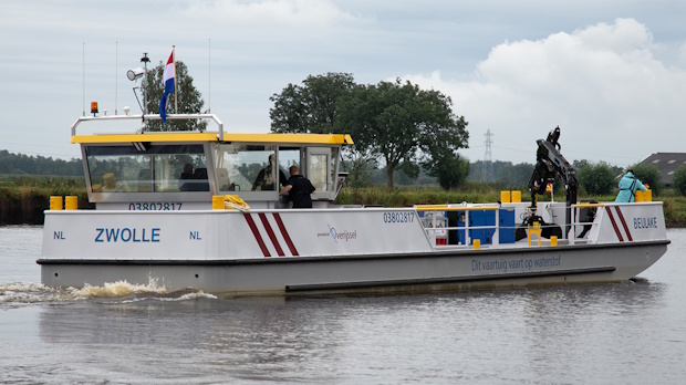 White passenger ferry with a yellow upper deck cruising on a calm river; Zwolle labeled on the hull, Dutch flag flying, passengers on board with trees in the background.