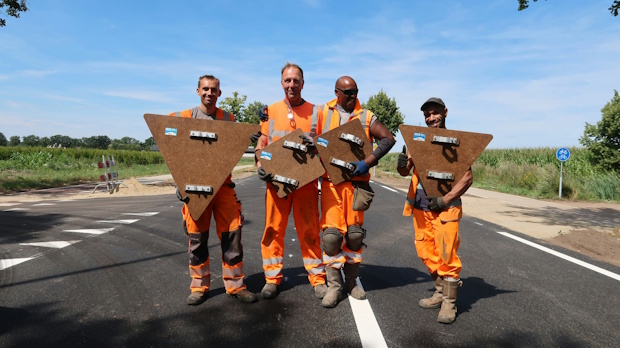 Four road workers in orange safety gear pose on a paved road, each holding a large metal road plate panel in front of them.