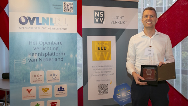 Smiling man in a white shirt at a trade show booth, standing beside banners for OVNL and NSNV, holding an award plaque and folder.