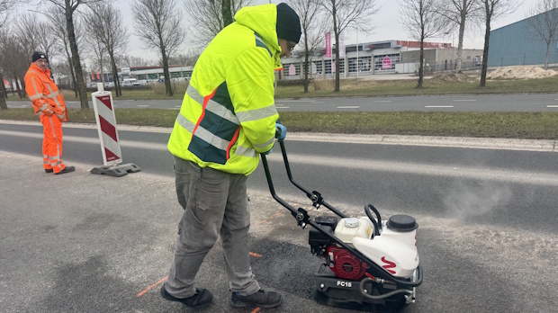 Worker in a neon high-visibility jacket operates a plate compactor on a road, with a second worker in orange nearby and buildings in the background.