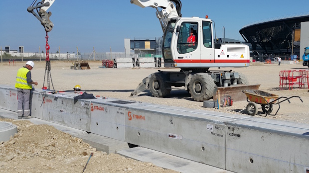 Construction site with a crane lifting a concrete beam, workers in safety vests, and a large excavator nearby.