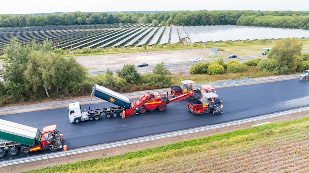 Aerial view of road construction: trucks and asphalt paver laying new road, with a roller compactor nearby and solar farms in the background.