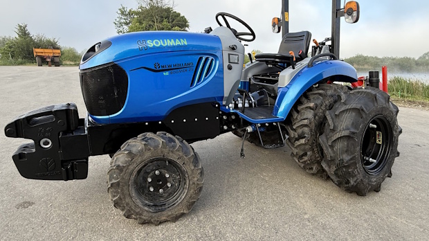 Side view of a blue compact utility tractor with large rear tires and a front hitch parked on a paved surface.