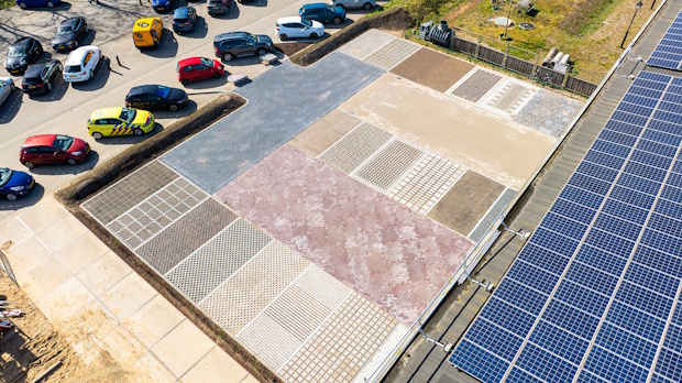 Aerial view of a parking lot with colorful cars at top left and a large patterned concrete area; on the right edge are rows of blue solar panels.