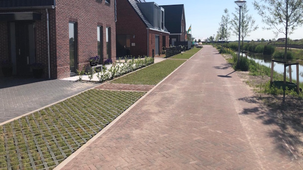 Sunlit suburban street with brick townhouses on the left, a pedestrian path, and a canal on the right.