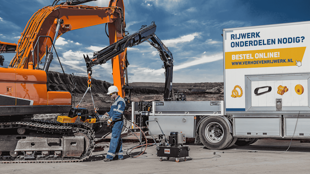 Construction site with an orange excavator and a worker in blue overalls and a white helmet inspecting hoses near a service truck.