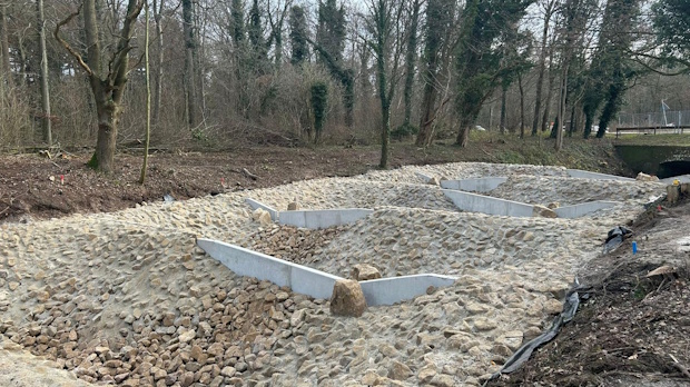 Construction site with rectangular stone-lined drainage basins and concrete walls in a trench along a wooded area, daytime