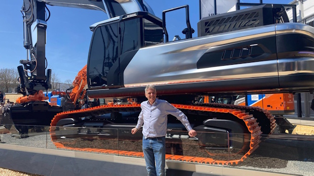Man in jeans and a button-down shirt poses in front of a large orange-tracked excavator on display outdoors.